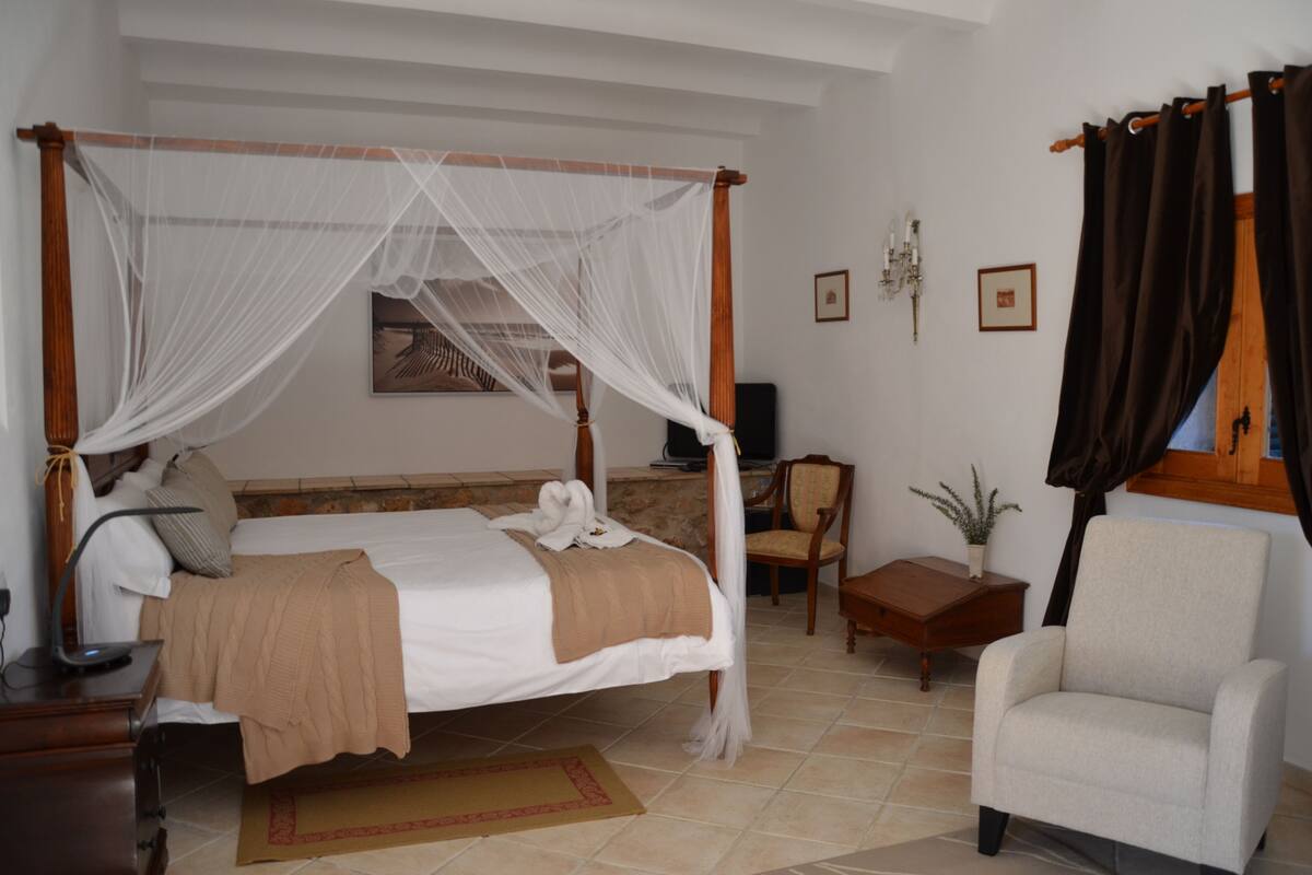 Un lit à baldaquin, une table de chevet avec une lampe, une chaise, une petite table avec une plante et un téléviseur fixé au mur.