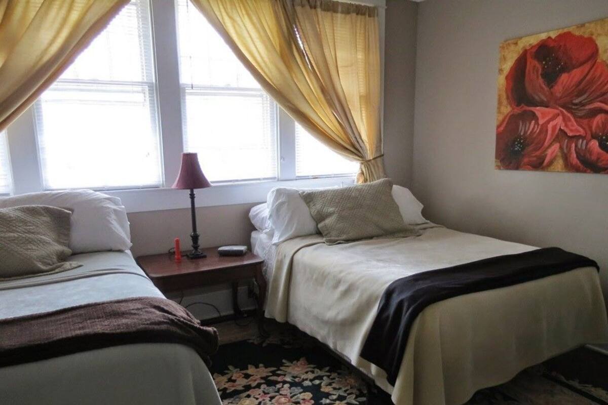 A room with two beds, a nightstand with a lamp, a window with curtains, and a painting on the wall.
