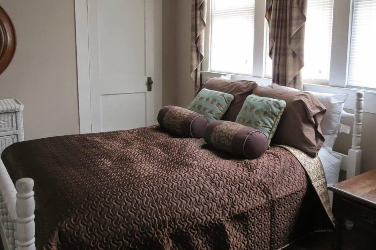 A bedroom with a patterned bedspread, two pillows, a nightstand, and a chair.