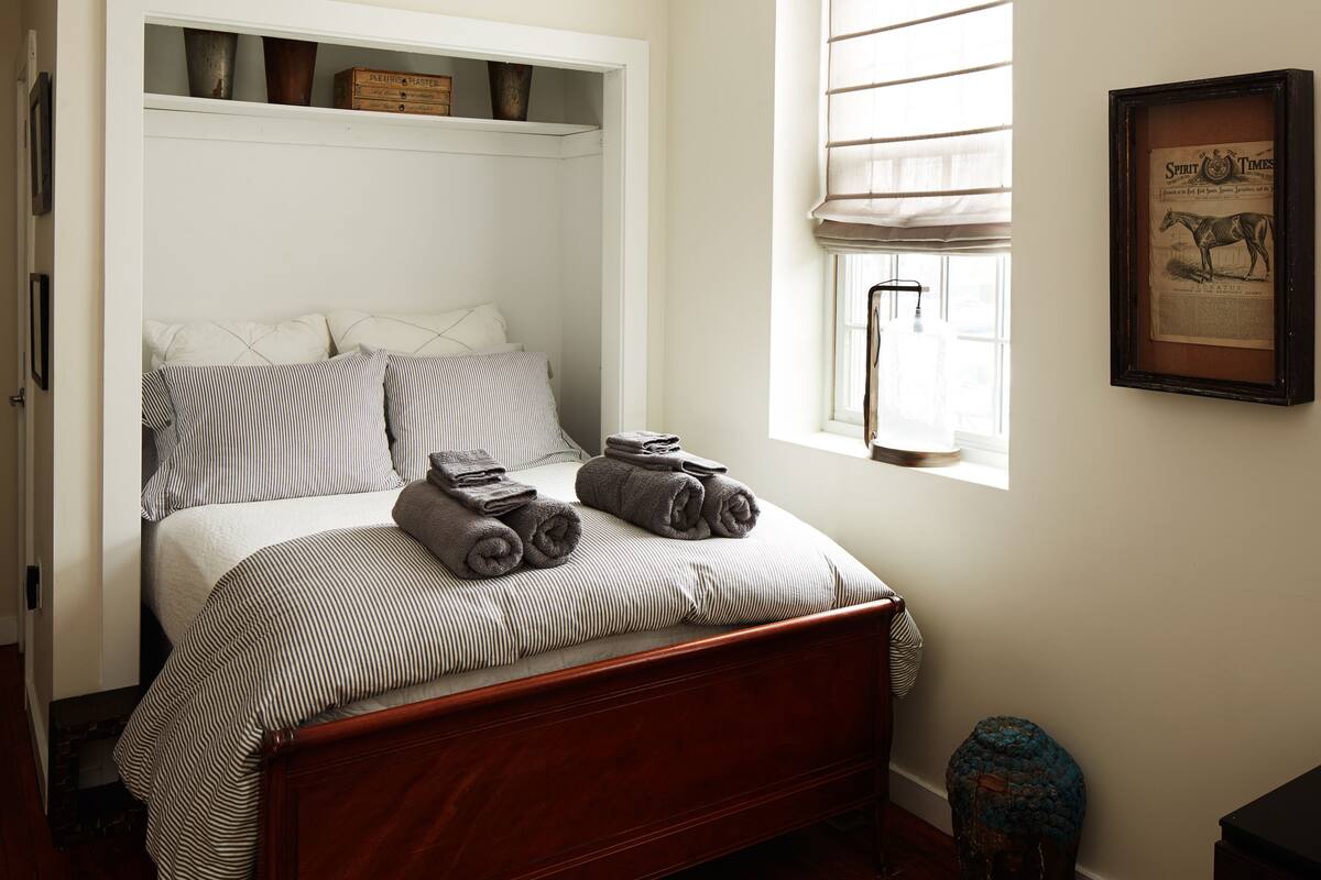 A neatly made bed with striped bedding and rolled towels, a window with blinds, and a framed newspaper on the wall.