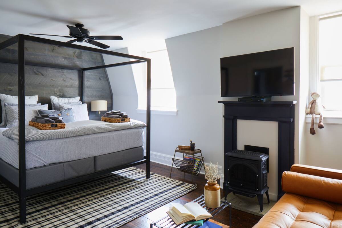 A bedroom with a four-poster bed, a television, a fireplace, and a window with curtains.