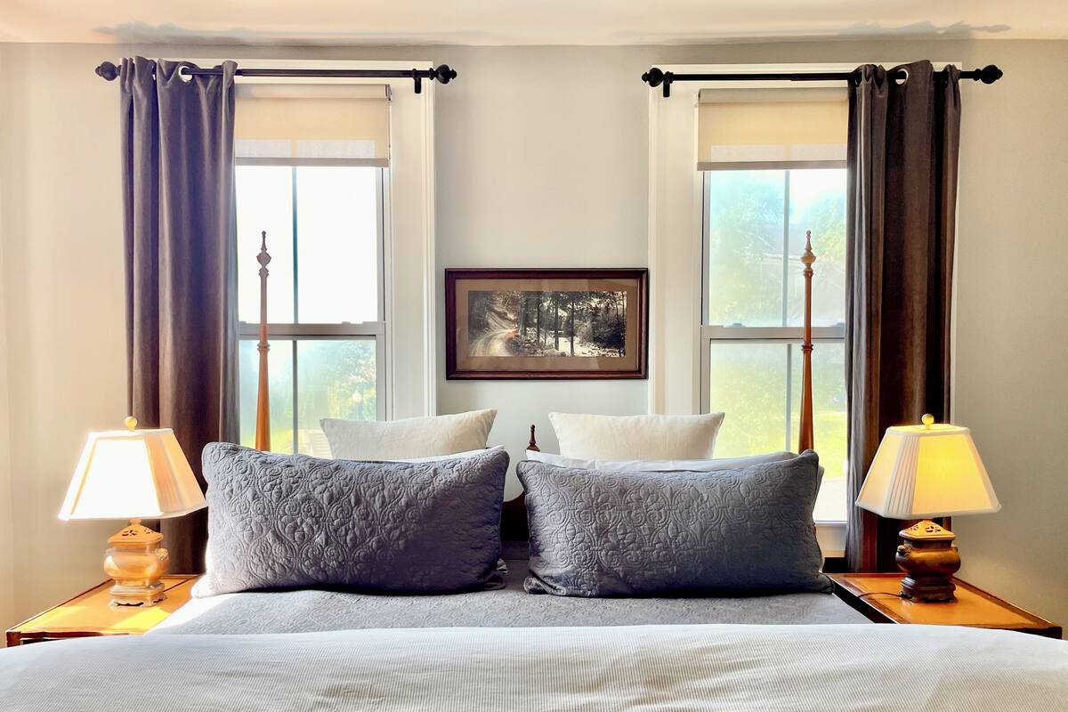 A bedroom with a large bed, two bedside lamps, a framed picture on the wall, and a window with curtains.