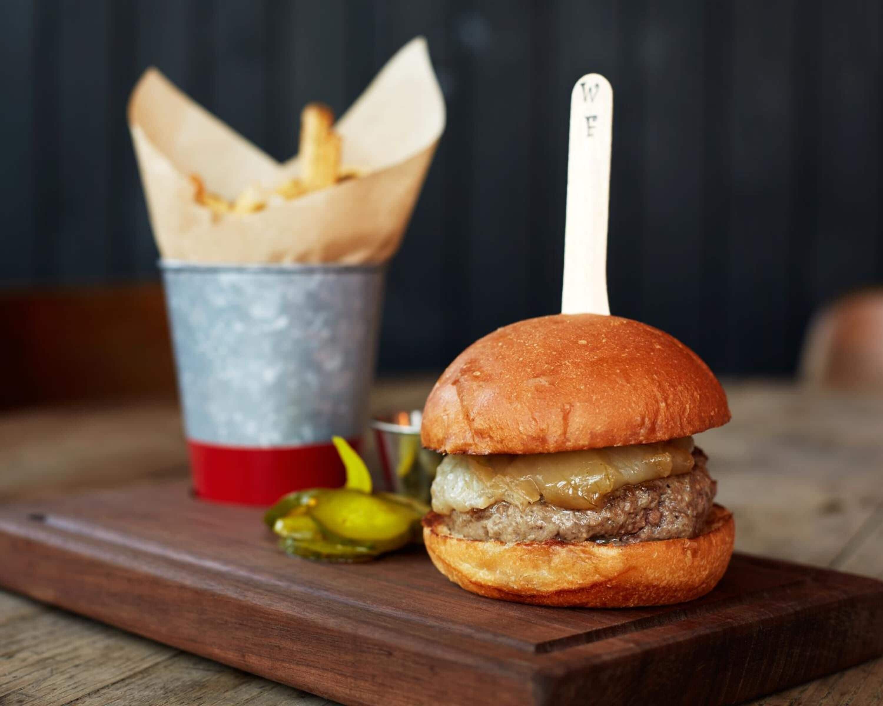 Una hamburguesa con un palillo sobre un pan, un cubo de papas fritas y pepinillos en una tabla de madera.