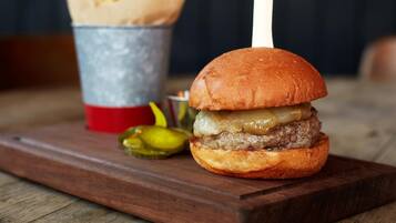 Una hamburguesa con un palillo sobre un pan, un cubo de papas fritas y pepinillos en una tabla de madera.