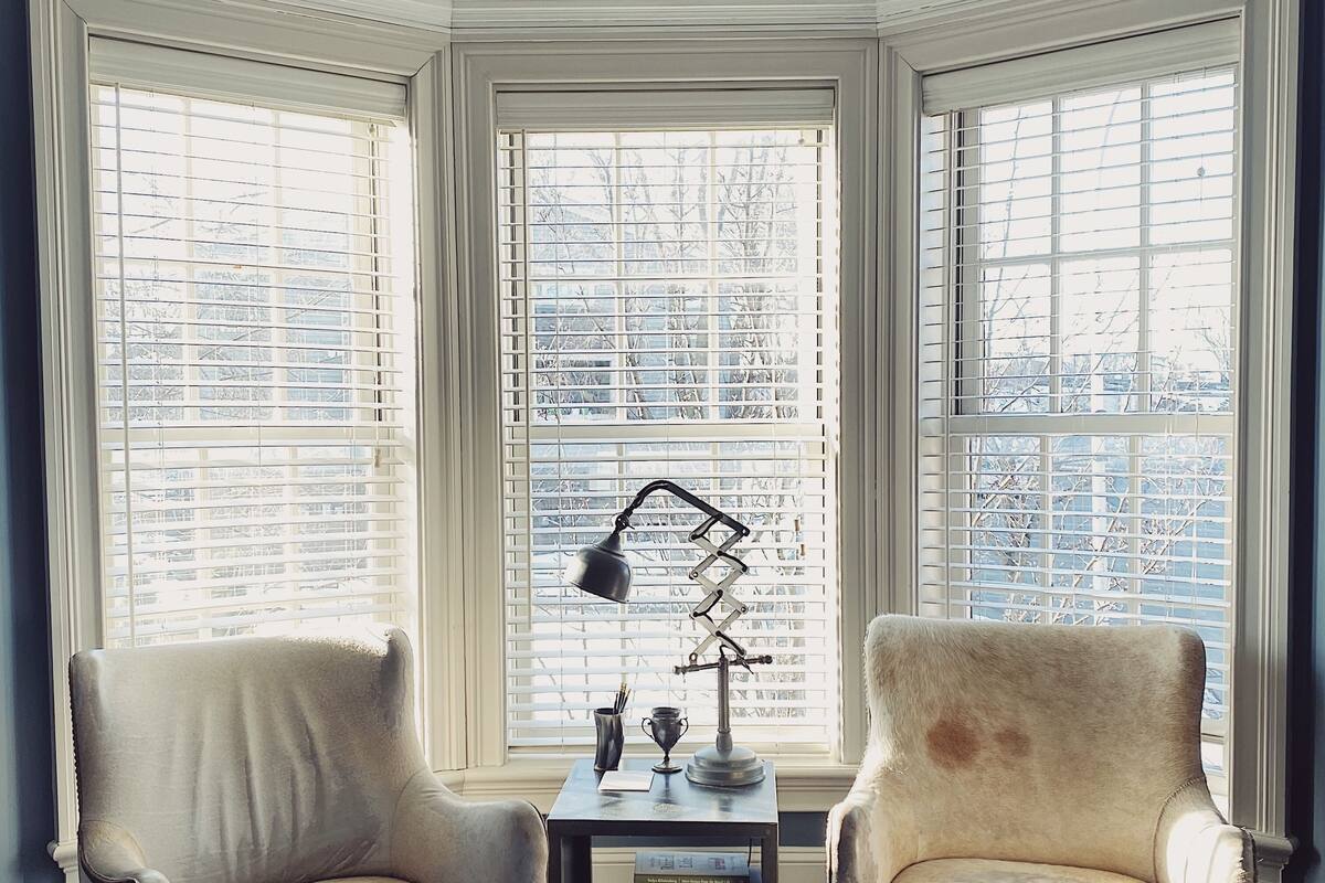 A room with two armchairs, a small table with a lamp, and a bookshelf. Large windows with blinds provide views of the outside.