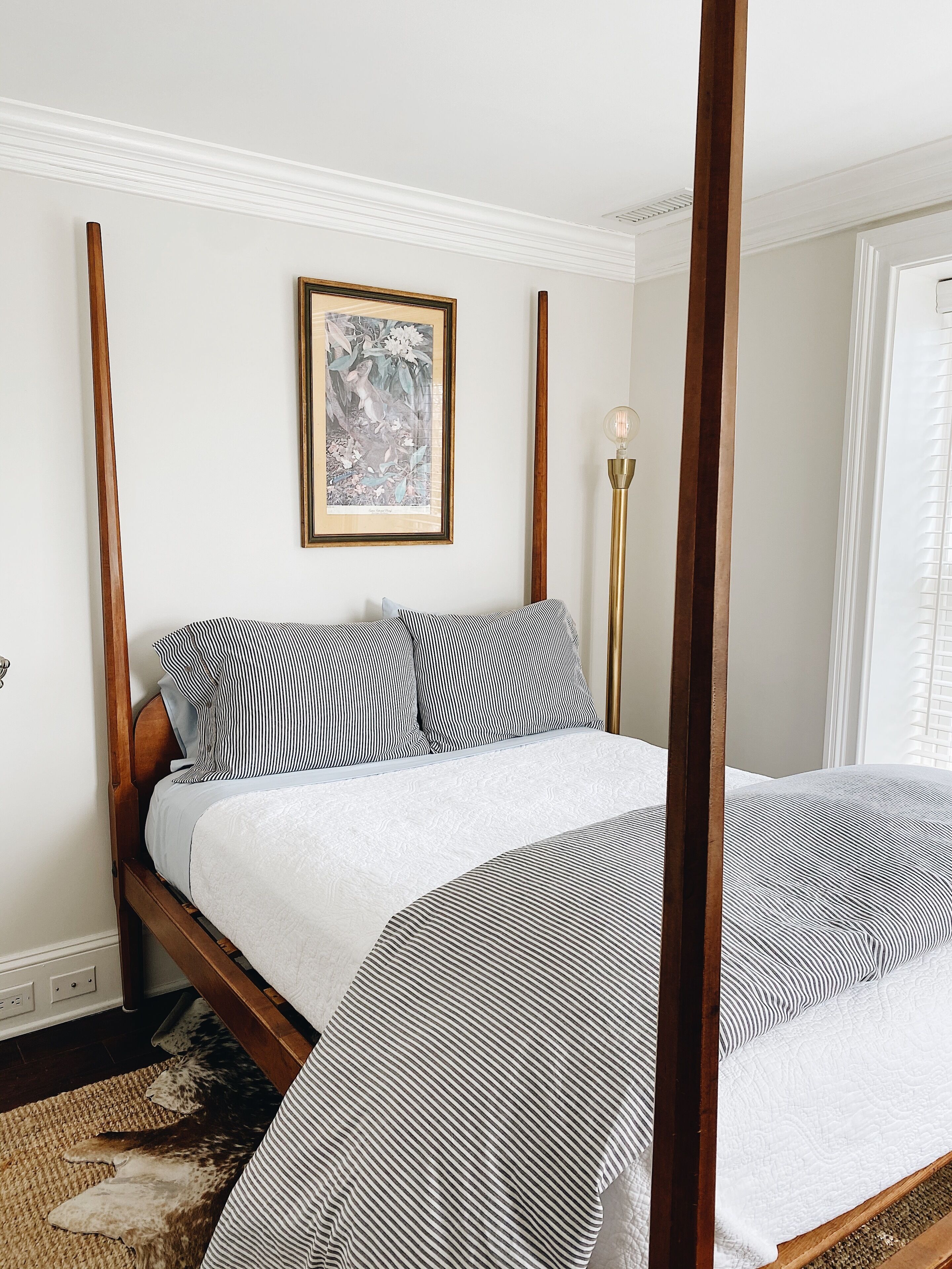 A four-poster bed with a white and black striped bedspread, a framed botanical print above the bed, and a tall floor lamp beside the bed.