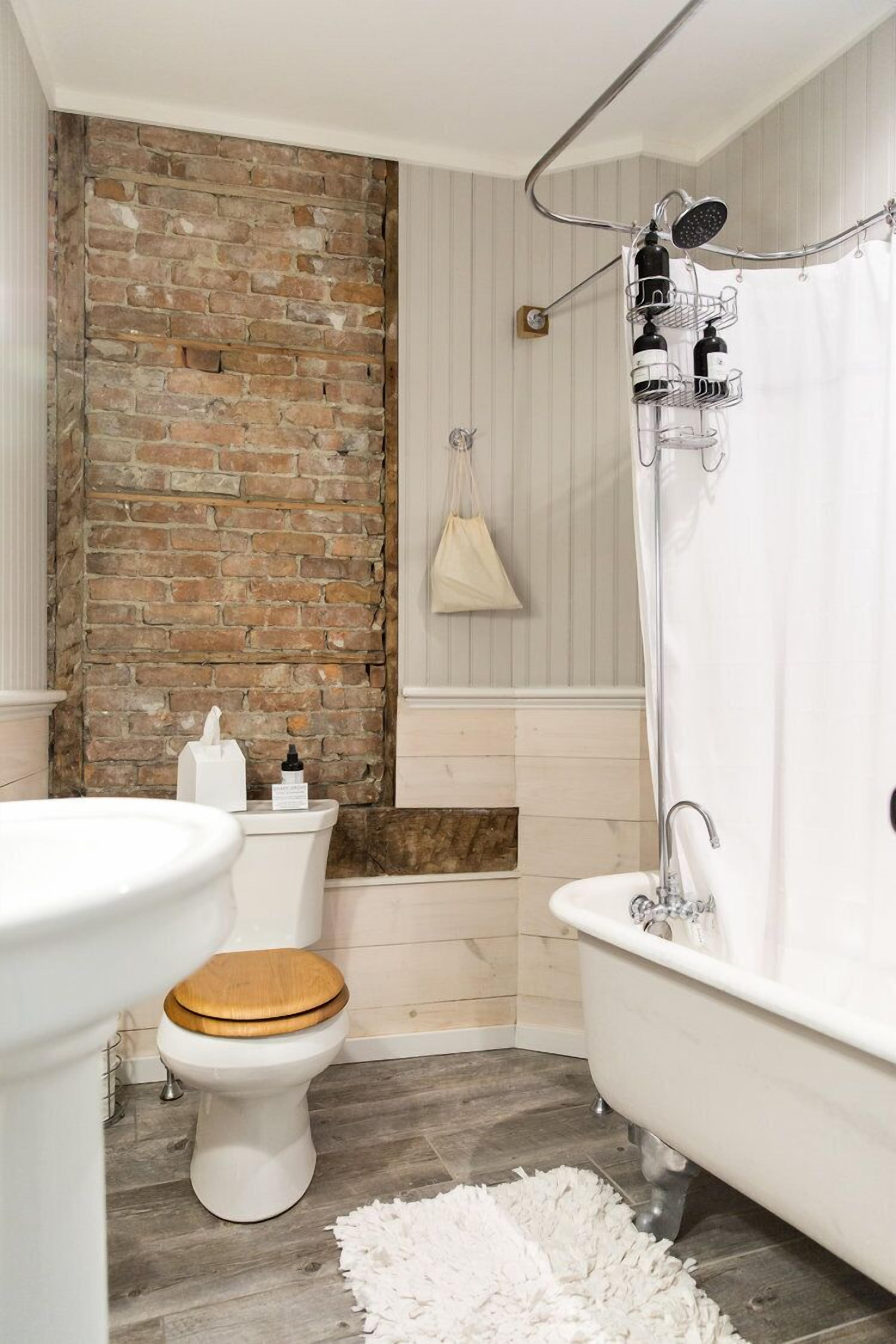 Un baño con pared de ladrillo, inodoro blanco, tapa de madera para el inodoro, lavamanos blanco, bañera blanca, ducha y cortina de ducha blanca.