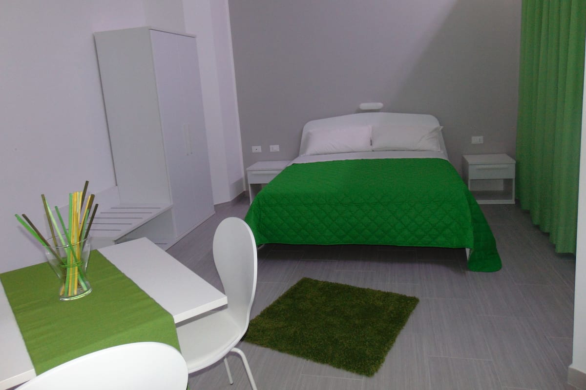 A bedroom with a bed, a desk, a chair, and a small table with a green tablecloth.