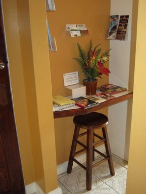 A small office area with a wooden desk, a stool, a potted plant, and various informational posters on the wall.