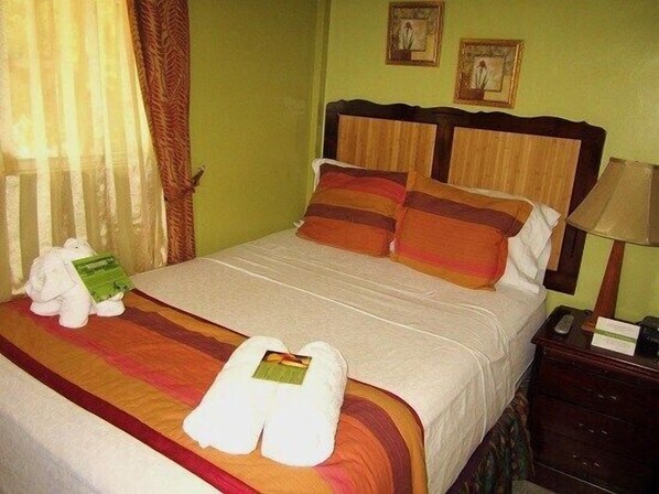 A neatly made bed with a striped bedspread, two pillows, and a stuffed animal holding a book. There is a pair of white slippers on the bed and a lamp on a bedside table.