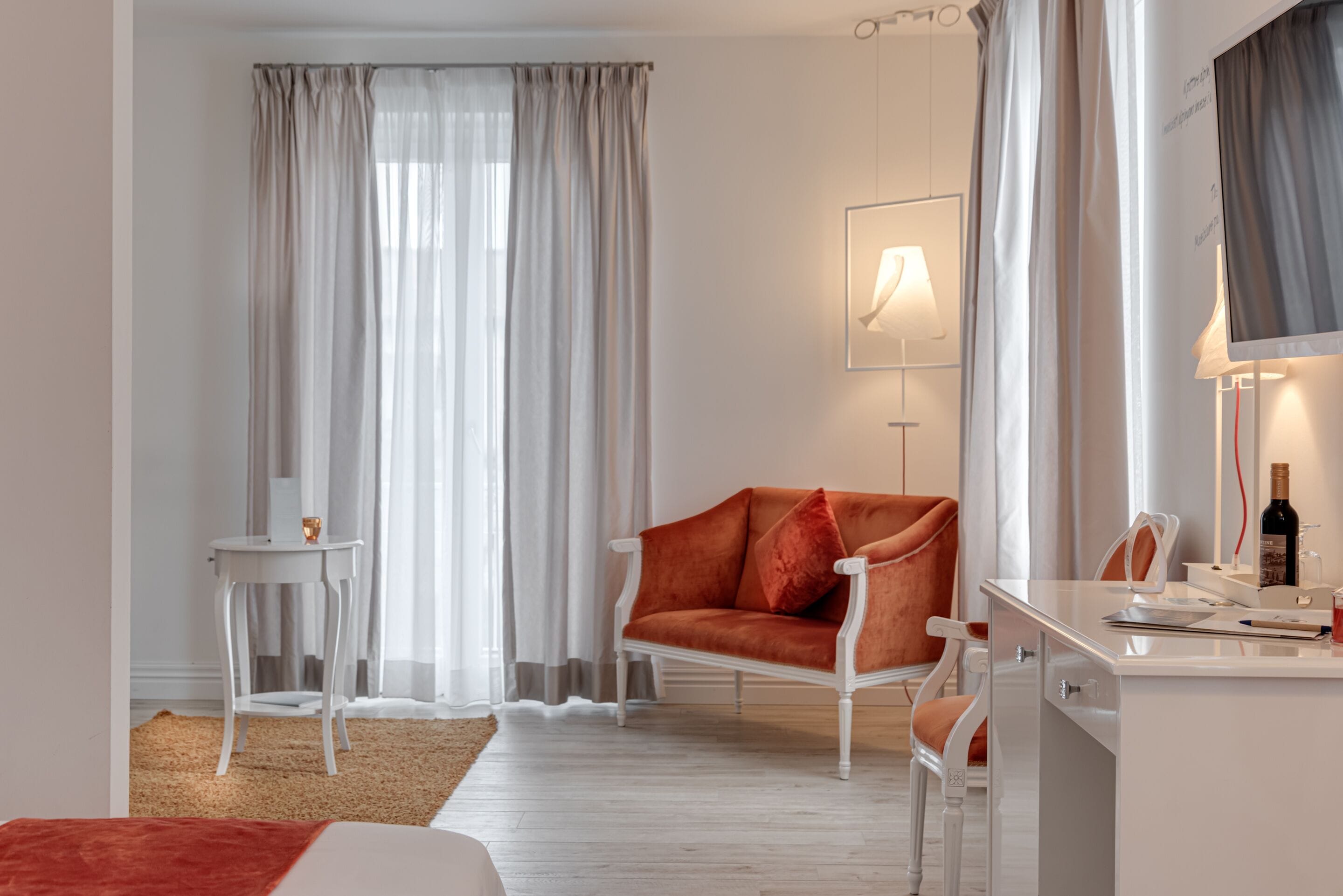 A modern hotel room with a white sofa, a small side table, a desk with a chair, and a large window with sheer curtains.