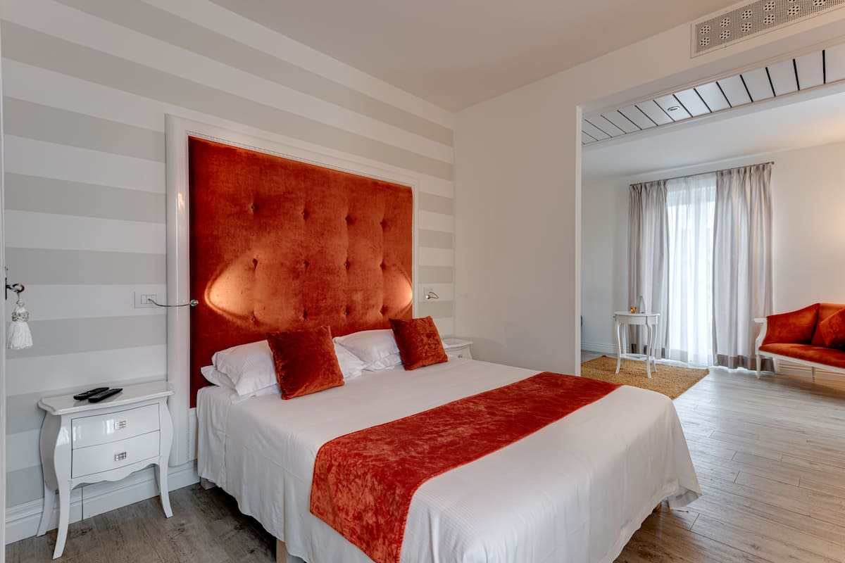 Una habitación de hotel moderna con una cama grande, un cabecero tapizado en rojo, ropa de cama blanca, una mesita de noche pequeña con una lámpara y un sofá rojo en la esquina.