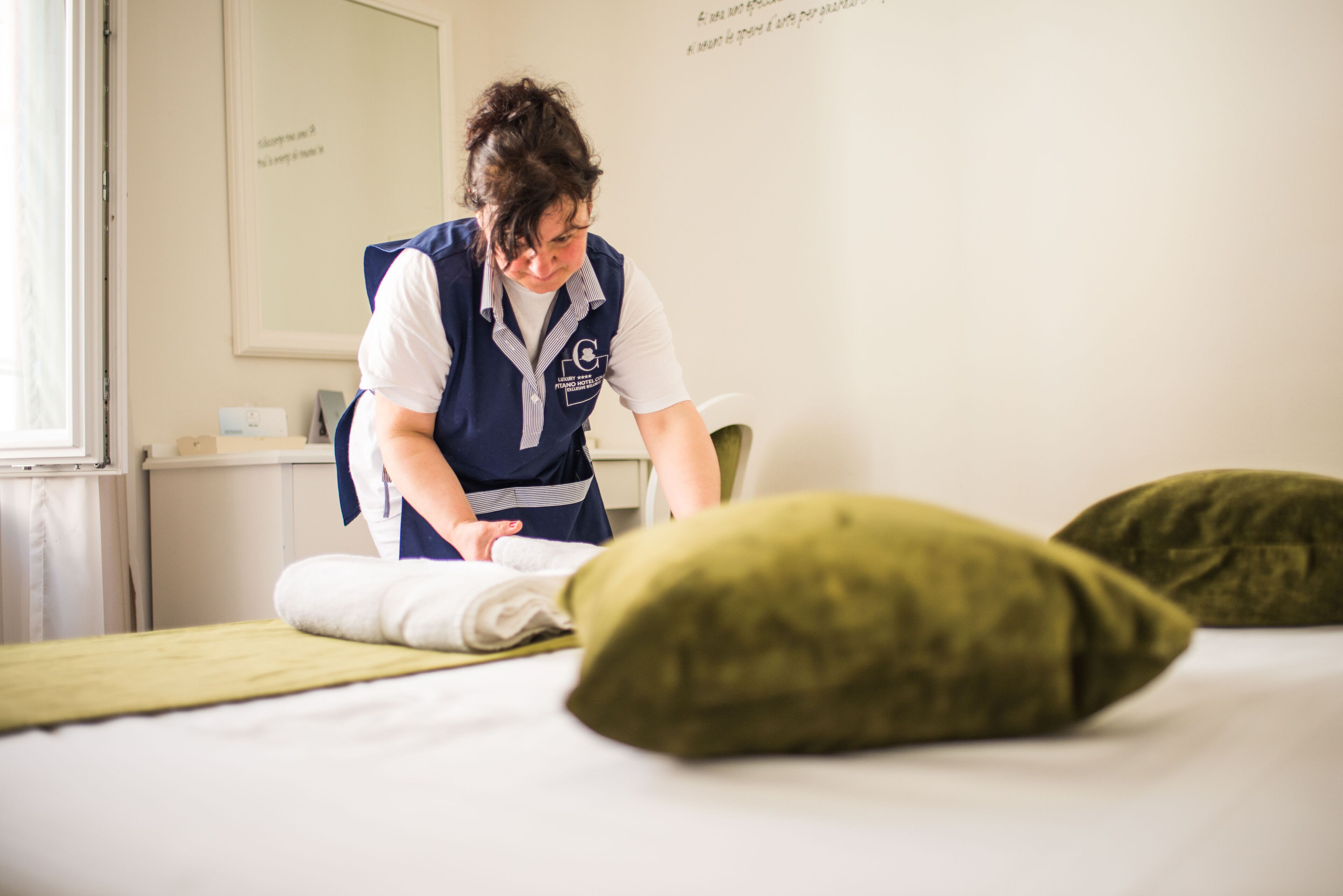 A person in a uniform folding a bed in a room with a window and a framed text on the wall.
