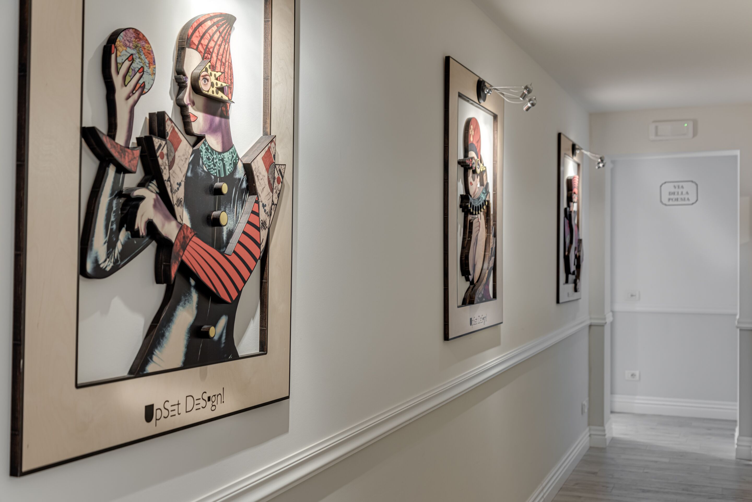 A hallway with framed artwork on the walls, a white door with a sign, and a light fixture above.