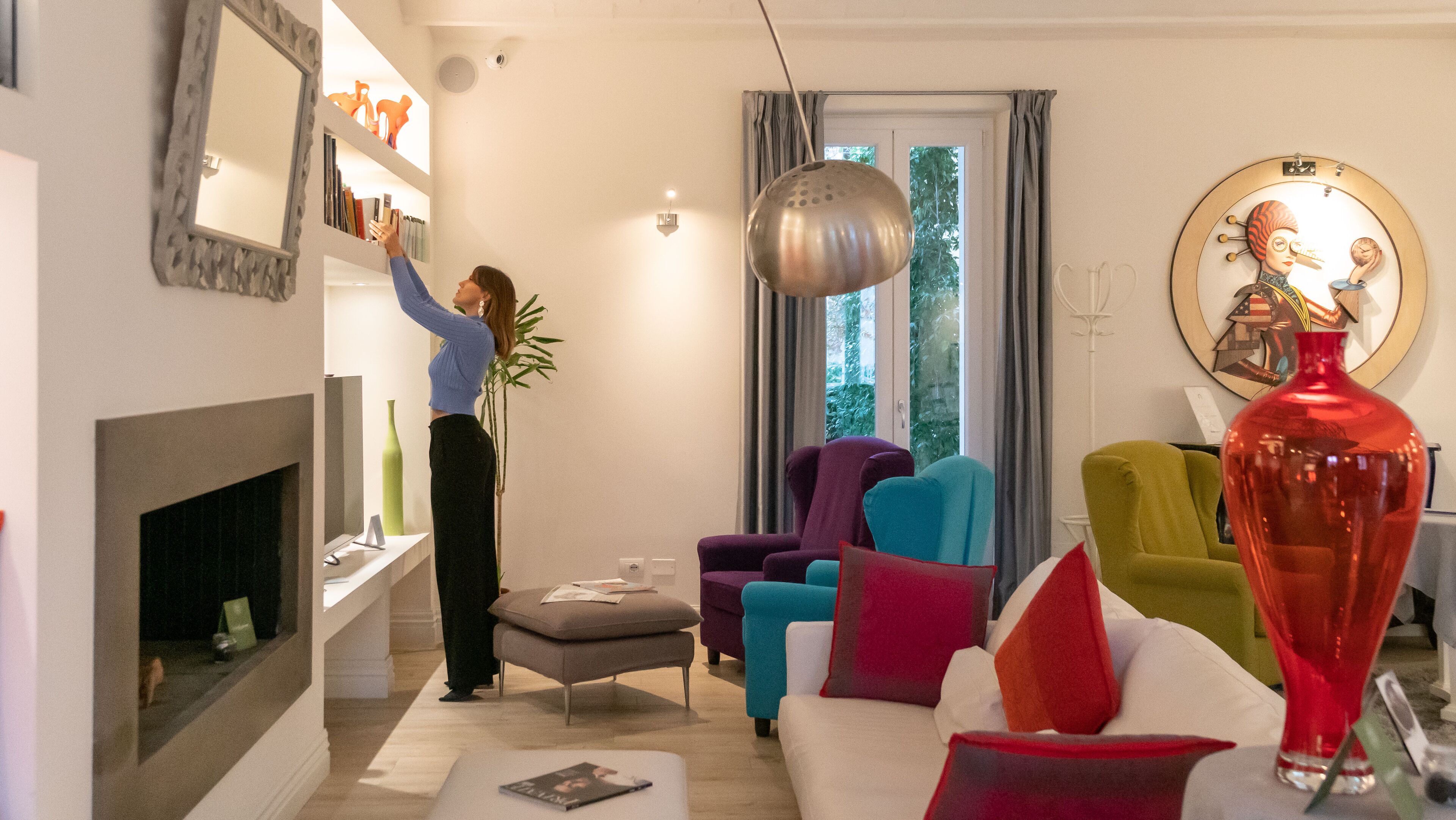 A living room with a fireplace, colorful sofas, a large red vase, and a person reaching for items on a shelf.
