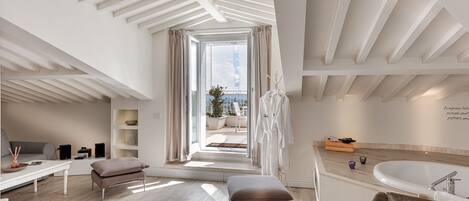 A modern hotel room with a large window, a bathtub, a seating area, and a view of the outdoors.