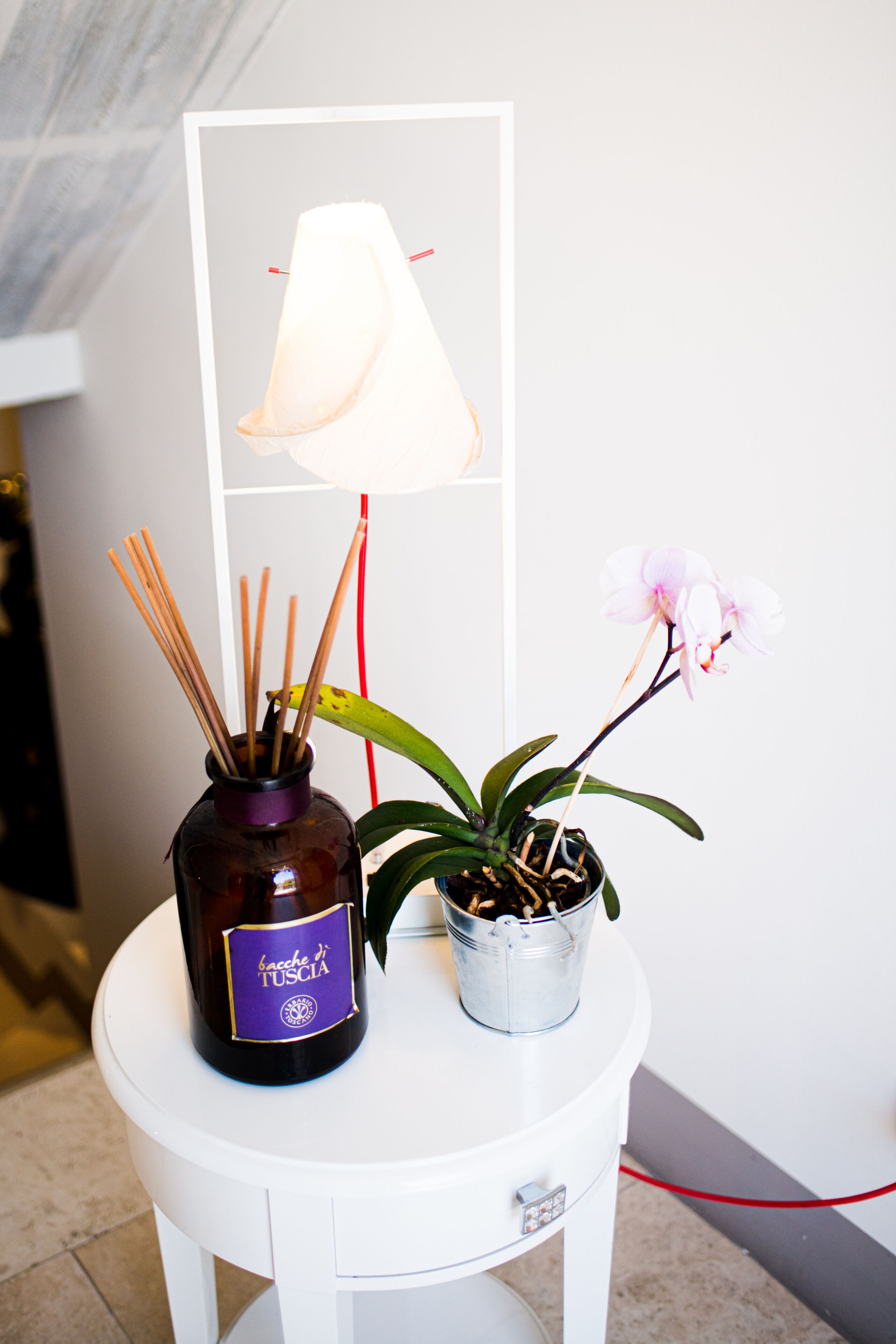 A small white table with a bottle of Bacche di Toscana, a potted plant, and a lamp.