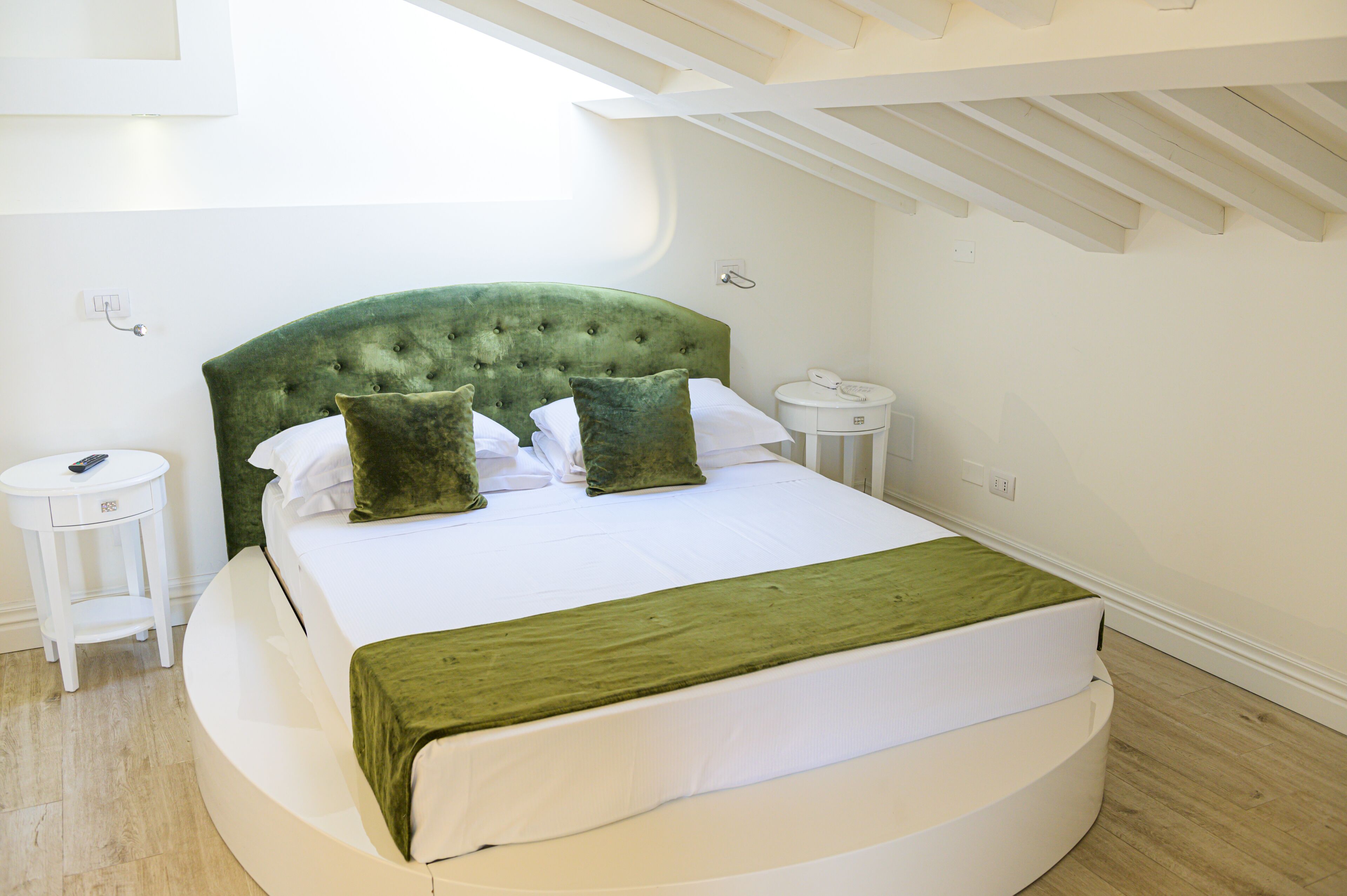 A neatly arranged bedroom with a green upholstered headboard, a round bed, white bedding, and a small bedside table with a remote control.