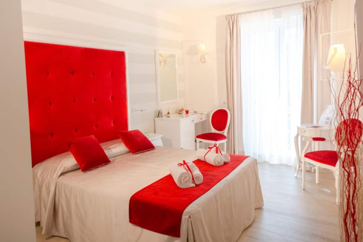 Una habitación de hotel con cabecera roja, cama con ropa de cama blanca y manta roja, además de un escritorio y una silla.