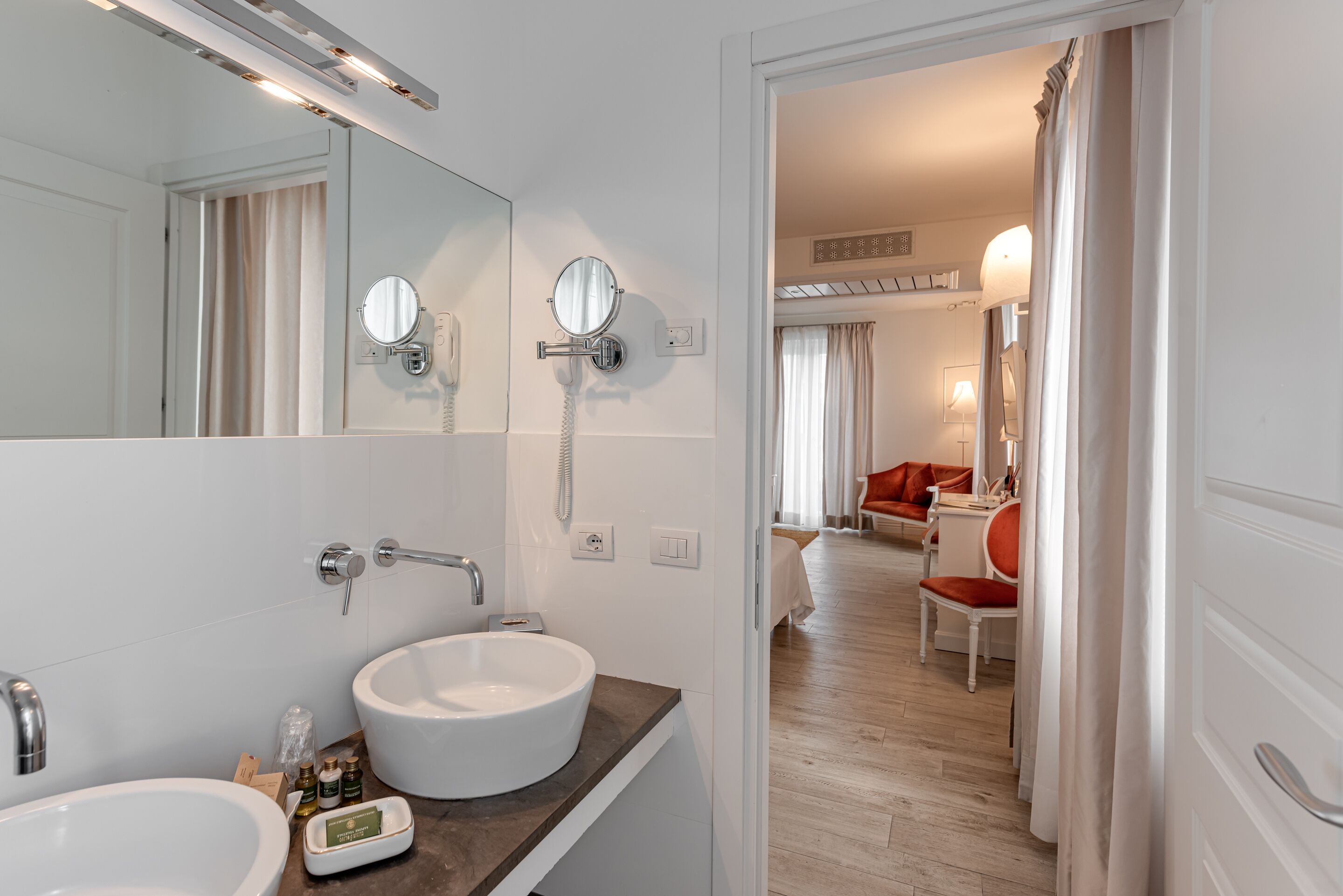 Um banheiro moderno com duas pias, um espelho grande e um telefone. O cômodo possui piso de madeira e vista para uma sala de estar com uma poltrona vermelha e uma mesa.