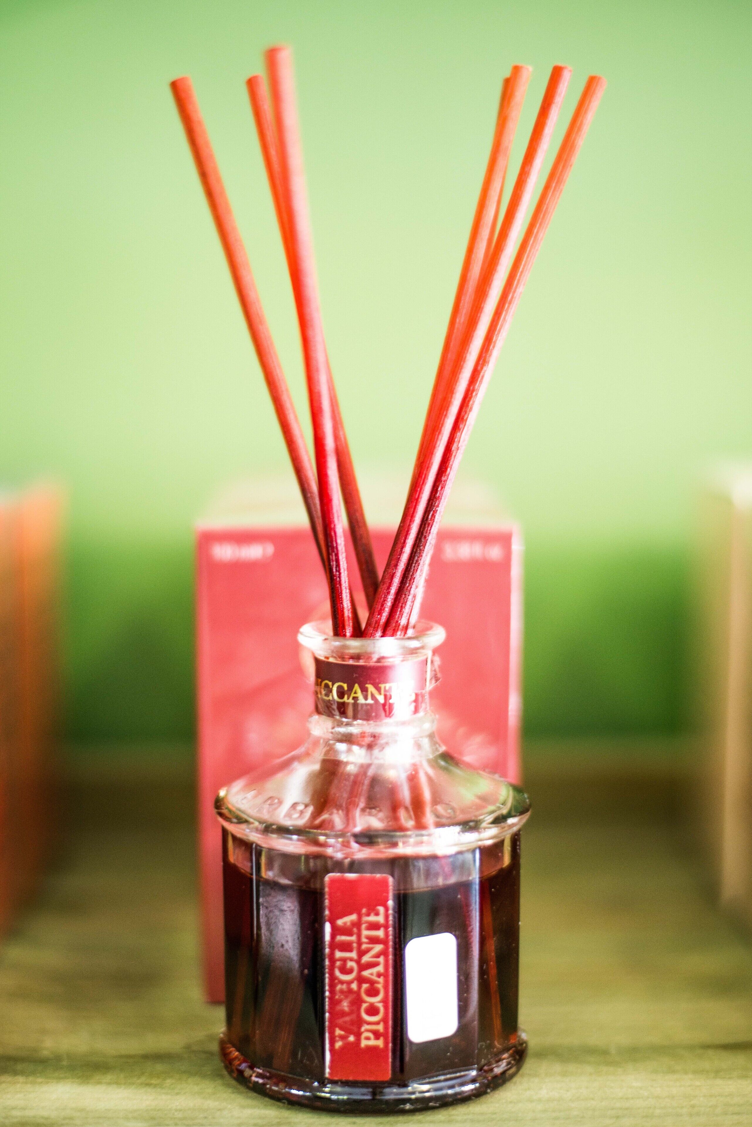A glass bottle with red reed sticks and a label reading "VAGLIA PICANTE".