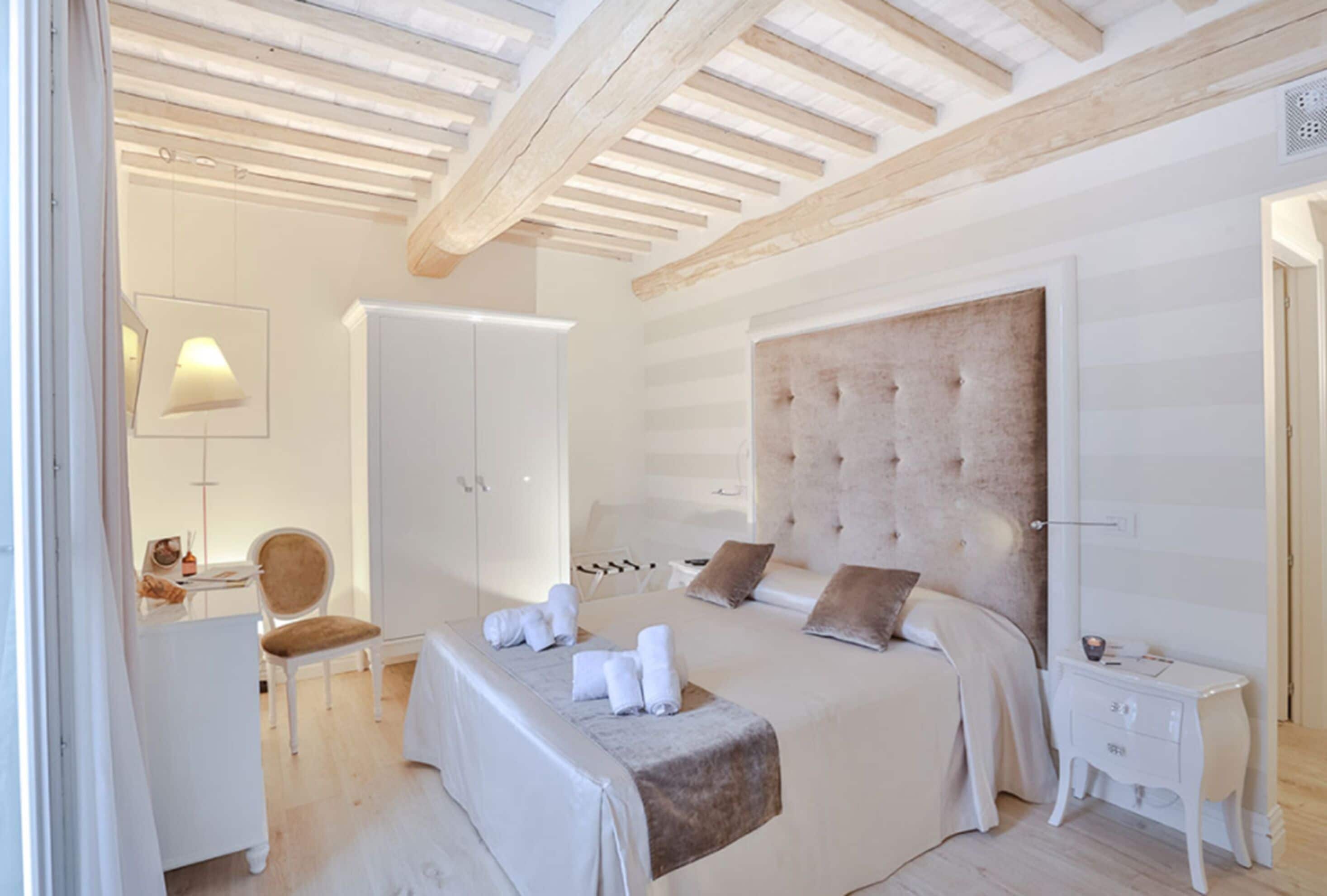 A bedroom with a large bed, a bedside table, a chair, a wardrobe, and a ceiling with wooden beams.