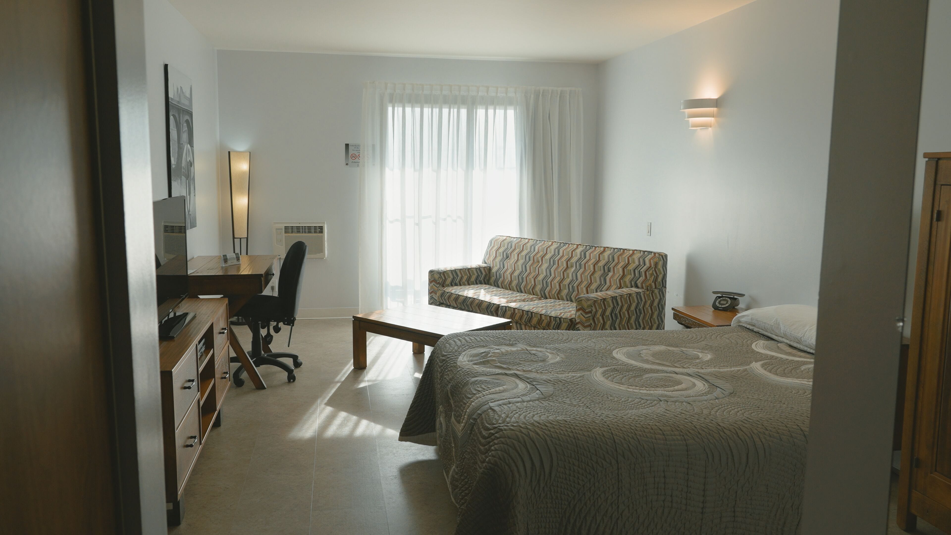 Habitación de hotel con cama, escritorio, silla, mesita, lámpara y ventana con cortinas.