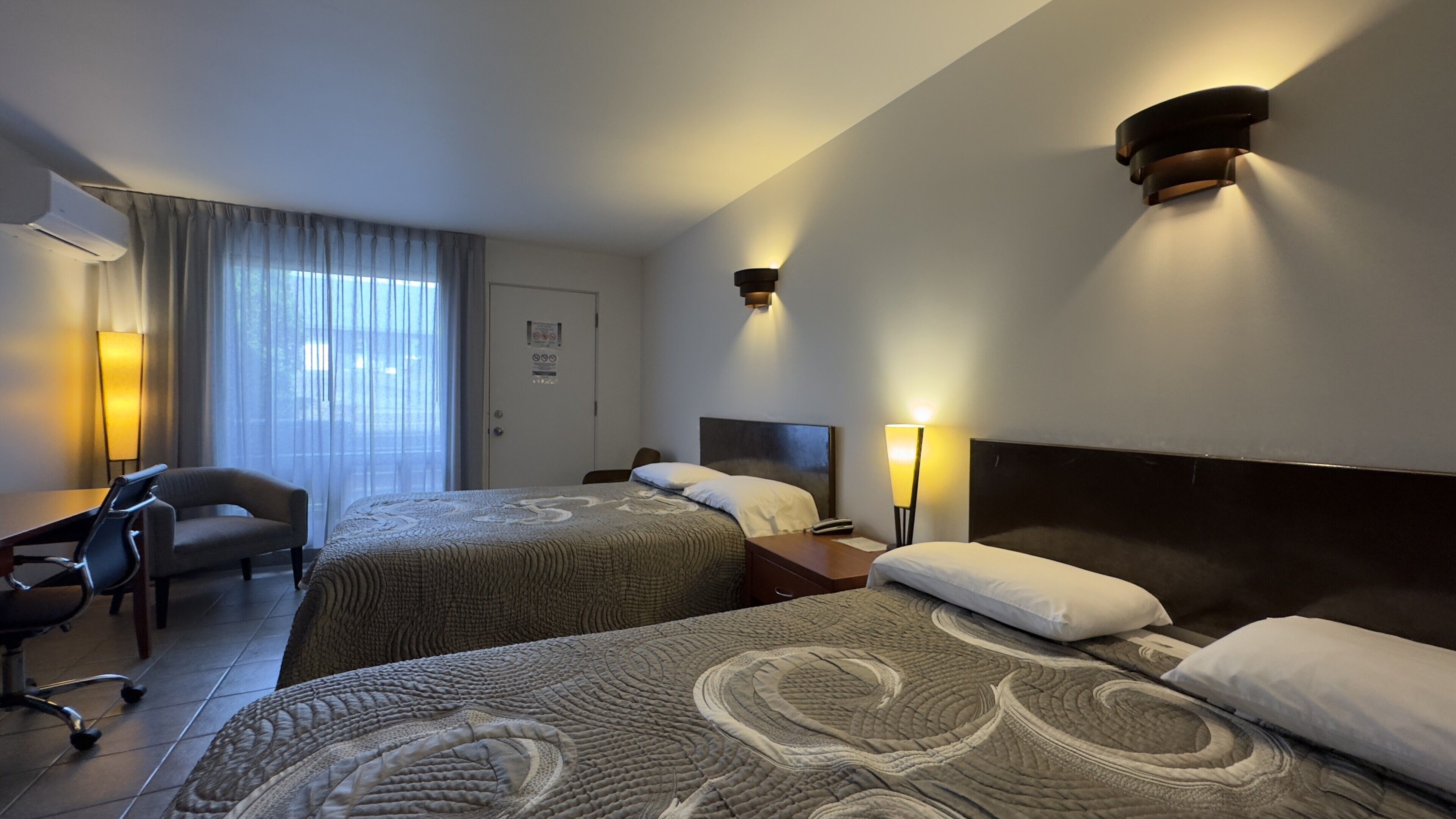 Une chambre d’hôtel avec deux lits, un bureau, une chaise et une grande fenêtre avec des rideaux.