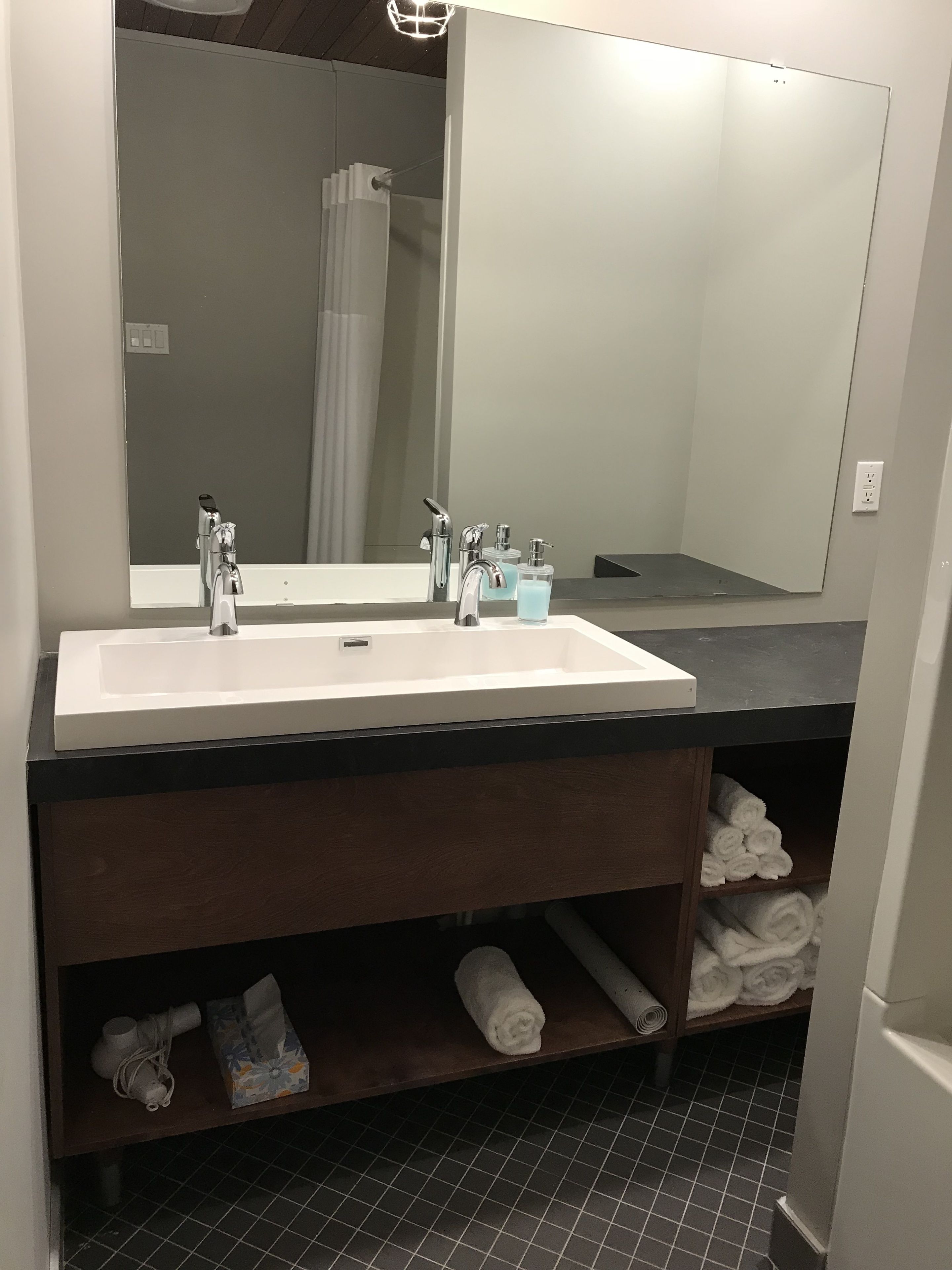 A bathroom with a white sink, two faucets, a mirror, and a towel shelf with towels.