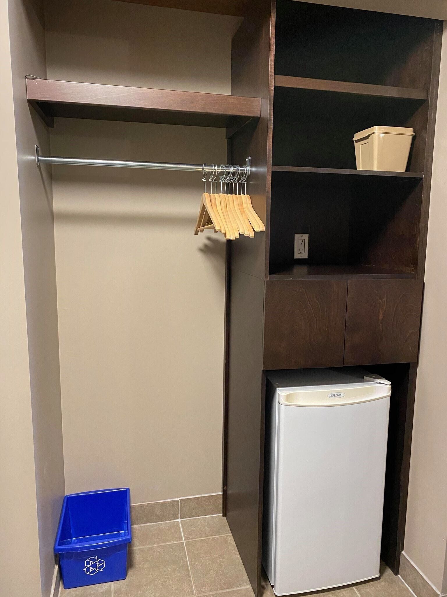 A compact kitchenette with a small refrigerator, a blue recycling bin, and a wooden clothes rack.