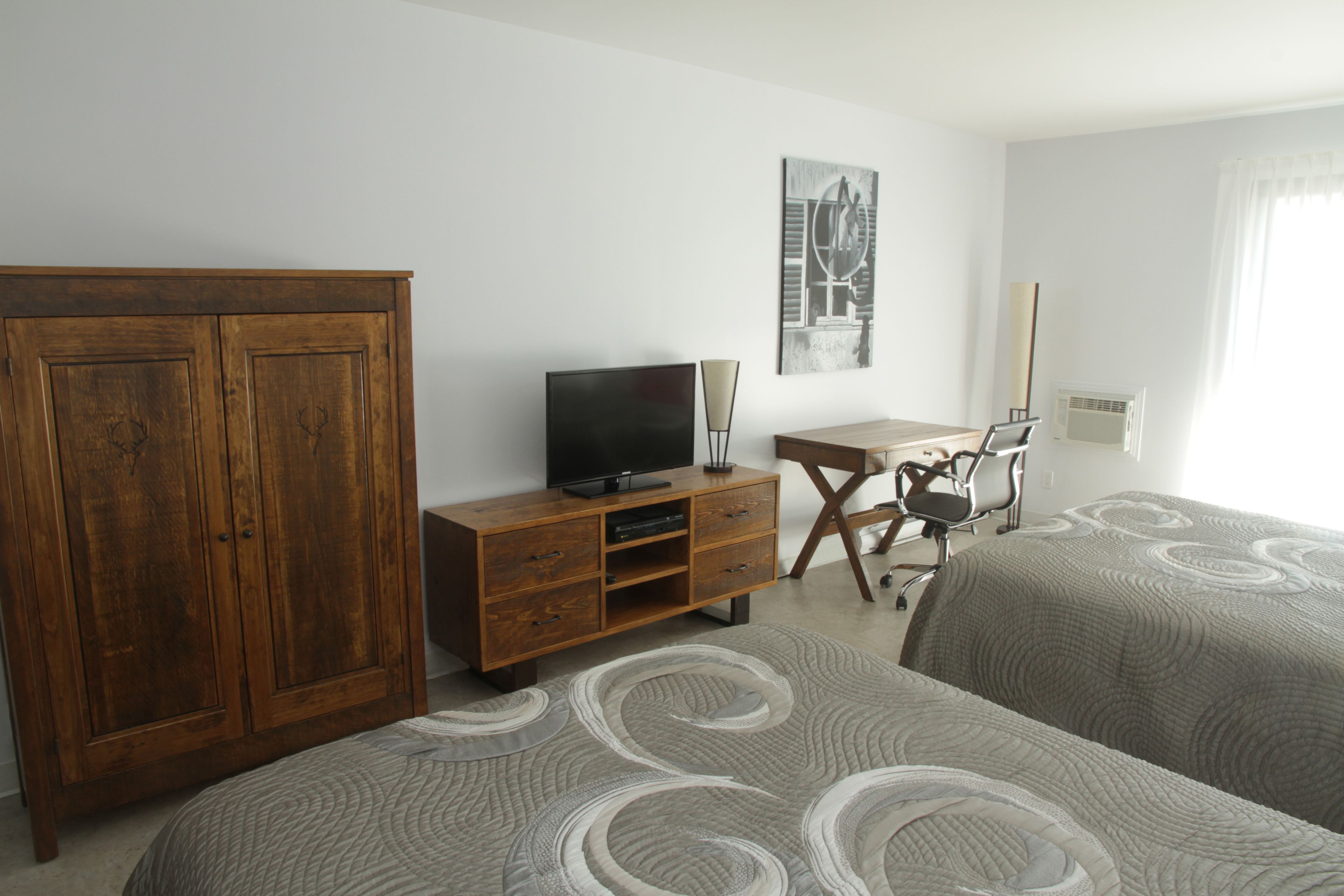 A bedroom with two beds, a wooden wardrobe, a desk with a chair, a television, and a painting on the wall.