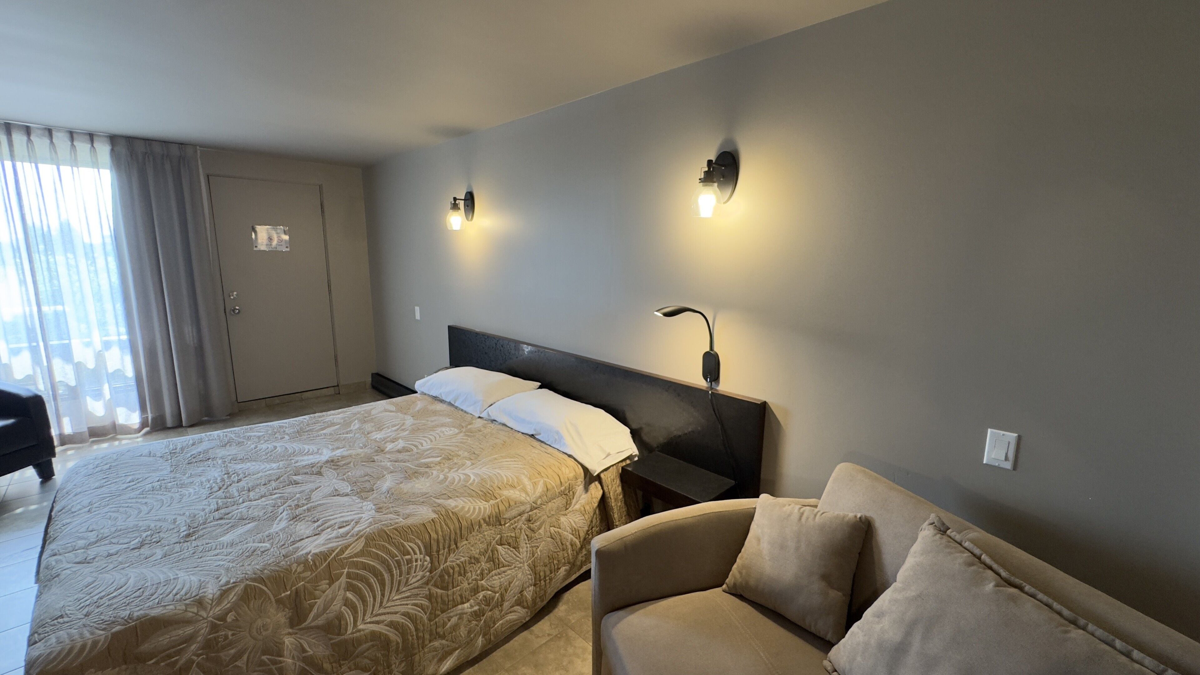 A hotel room with a large bed, a bedside table with a lamp, a sofa, and a door with a curtain.