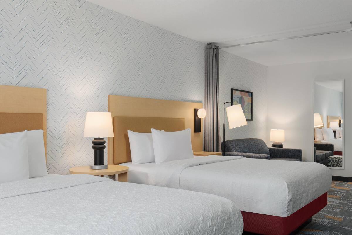 Ein Hotelzimmer mit zwei Betten, einem Kopfteil, einem Nachttisch, einer Lampe, einem Sessel und einem Spiegel, sowie einer Wand mit Muster.
