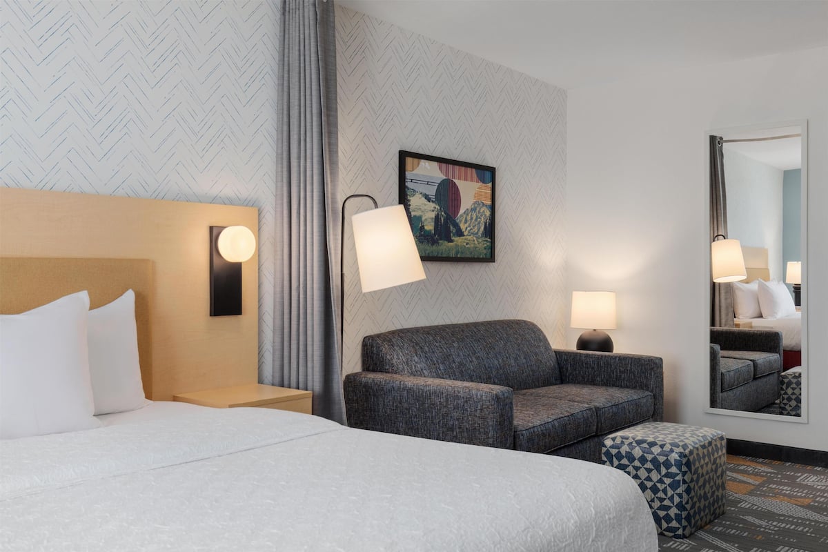 Ein Hotelzimmer mit Bett, Sofa, Sessel, Nachttisch, Lampe, Spiegel und Wand mit Fischgrätmuster.