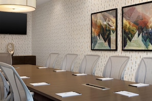 Une salle de conférence avec une longue table, six chaises, un téléviseur fixé au mur et deux tableaux abstraits encadrés.