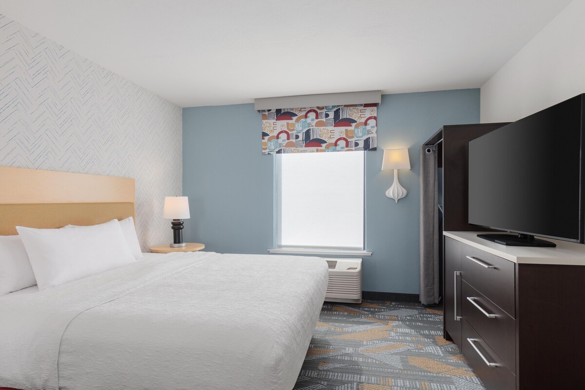 A hotel room with a bed, a television, a window with a patterned curtain, a lamp, and a patterned carpet.
