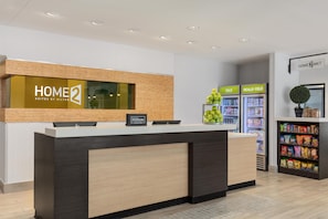 A hotel reception desk with a 'HOME2 Suites by Hilton' sign, a 'Hilton Honors' card, and a snack bar with 'COLD' and 'REALLY COLD' refrigerators. - Home2 Suites by Hilton Salt Lake City East (Salt Lake City)