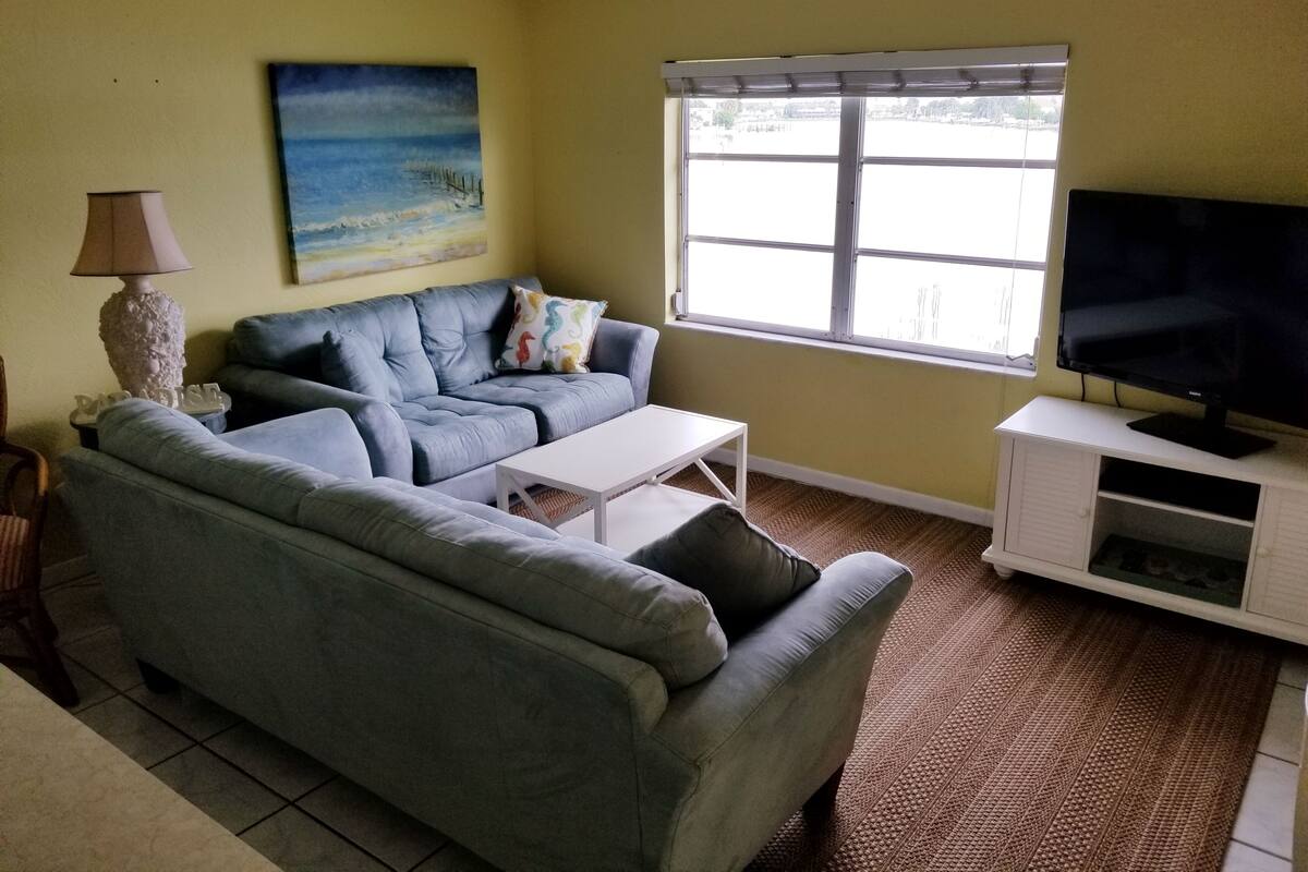 Ein Wohnzimmer mit grauem Sofa, einem weißen Couchtisch, einem Fernseher auf einem weißen Schrank, einem Gemälde an der Wand und einem Fenster mit Jalousien.