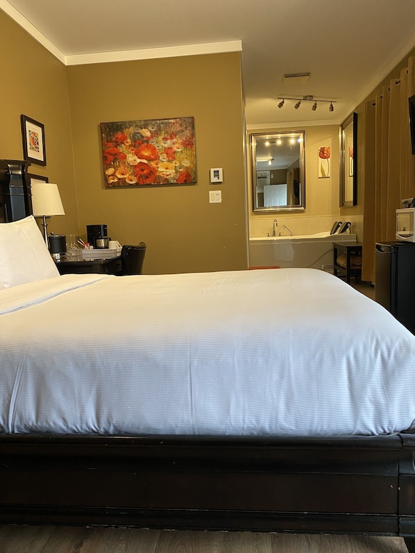 A hotel room with a large bed, a desk with a lamp, a painting on the wall, and a bathroom visible through an open door.