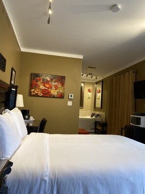 A hotel room with a large bed, a painting on the wall, a desk with a lamp, a coffee maker, and a microwave on a small table. - Motel des Cèdres (Sherbrooke)