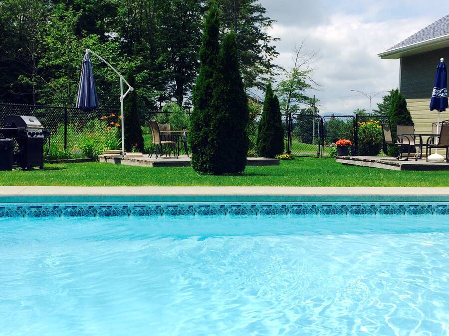 Ein Schwimmbecken mit gefliestem Rand, umgeben von einer Rasenfläche mit Grill, Stühlen und einem Zaun.