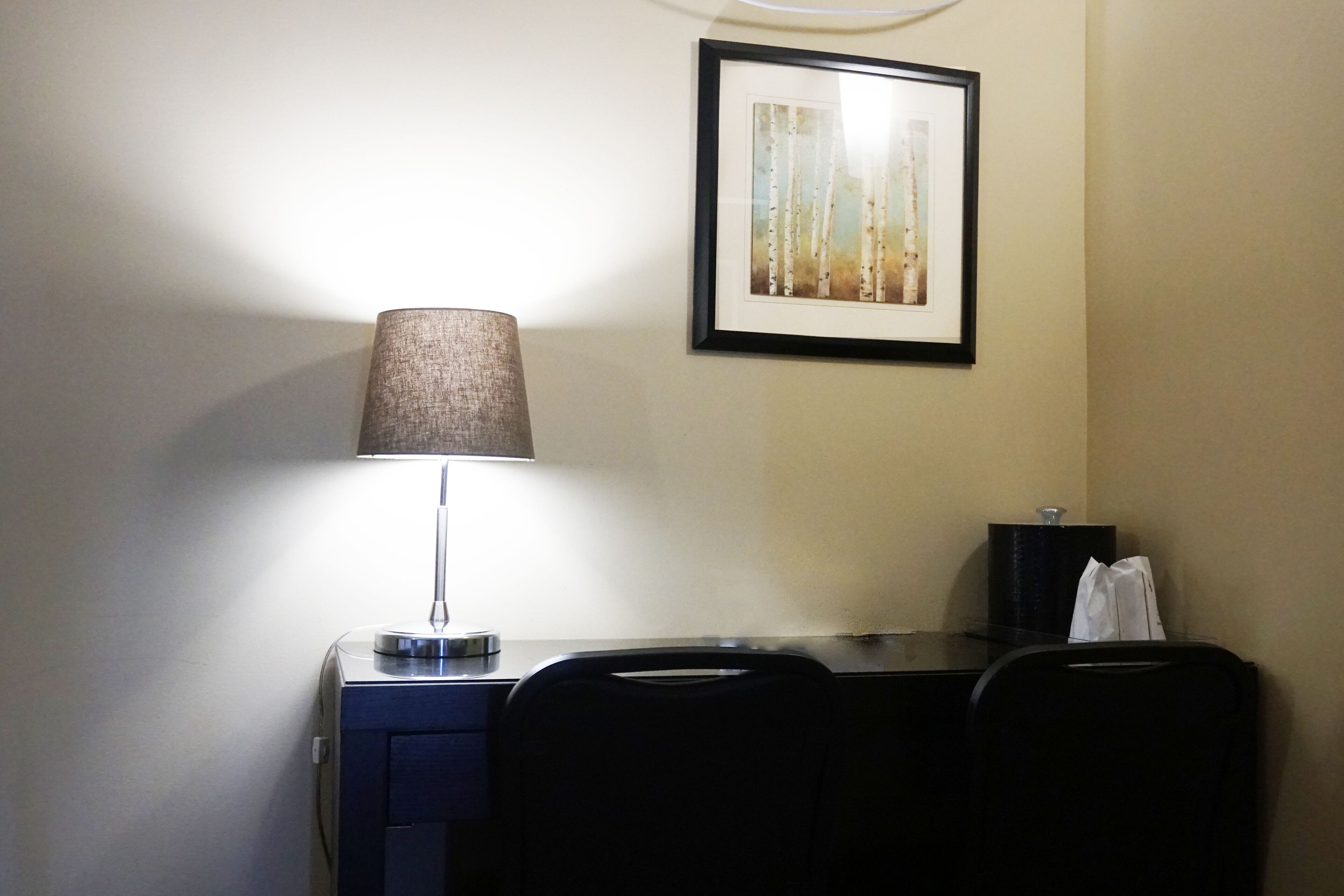 A desk with a lamp, a framed picture on the wall, and a black bag on the desk.