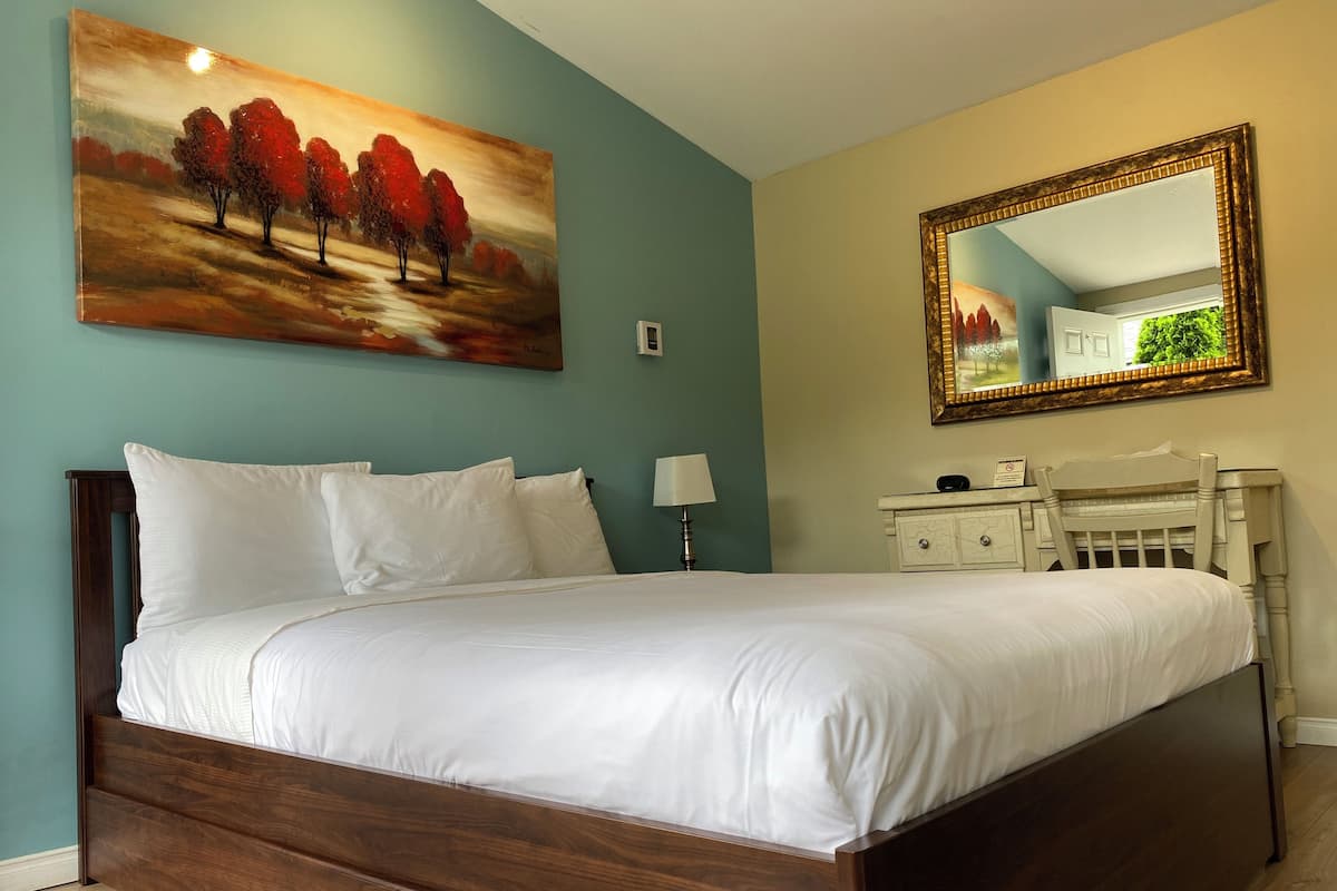 A neatly made bed with white linens in a room with teal walls, a large painting of trees, and a mirror with a gold frame.