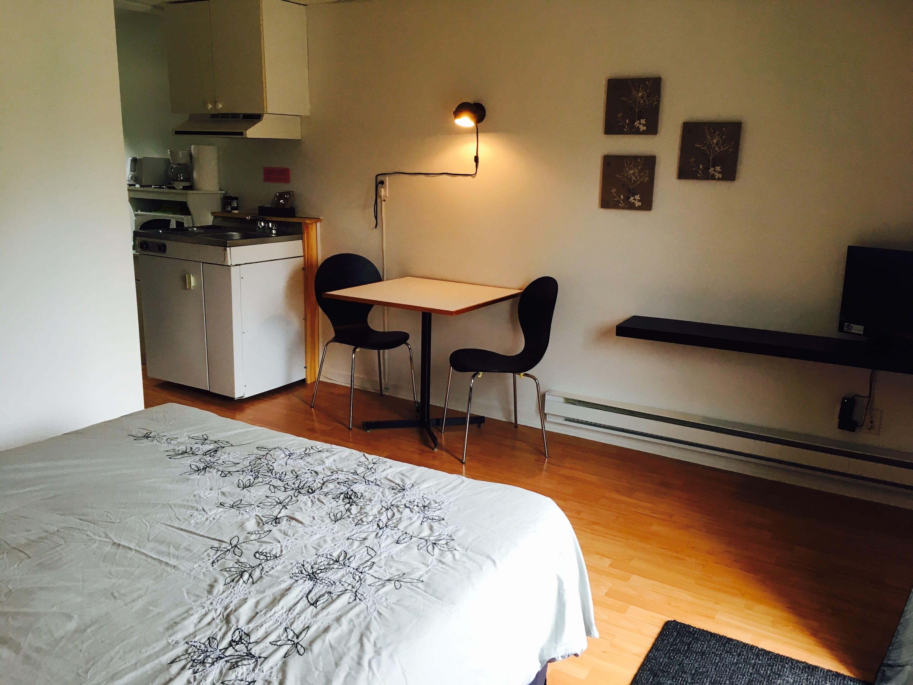 A compact bedroom with a bed, a small desk, two chairs, a kitchenette, and a TV shelf.