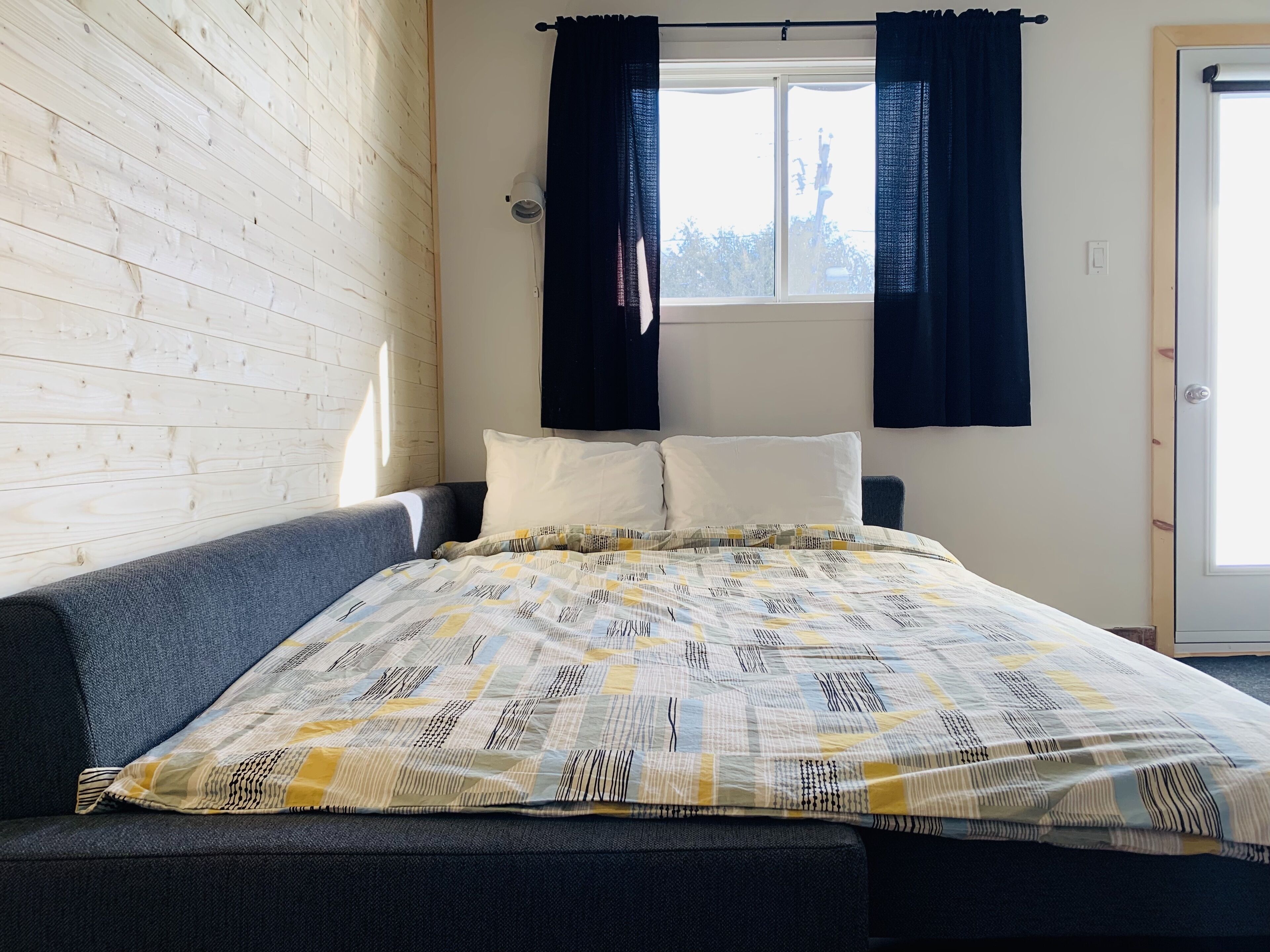 A bedroom with a bed, a grey sofa, a window with curtains, and a wooden wall.