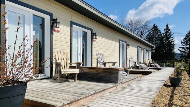 A wooden deck leading to a row of rooms with sliding glass doors, outdoor seating, and a view of trees and mountains. - Attitude Montagne (St-Adolphe-d'Howard)