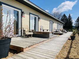 Una terraza de madera que conduce a una hilera de habitaciones con puertas correderas de vidrio, mobiliario exterior y vistas a árboles y montañas.
