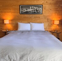Una cama con ropa de cama blanca, dos almohadas blancas y una fotografía enmarcada de montañas en la pared.