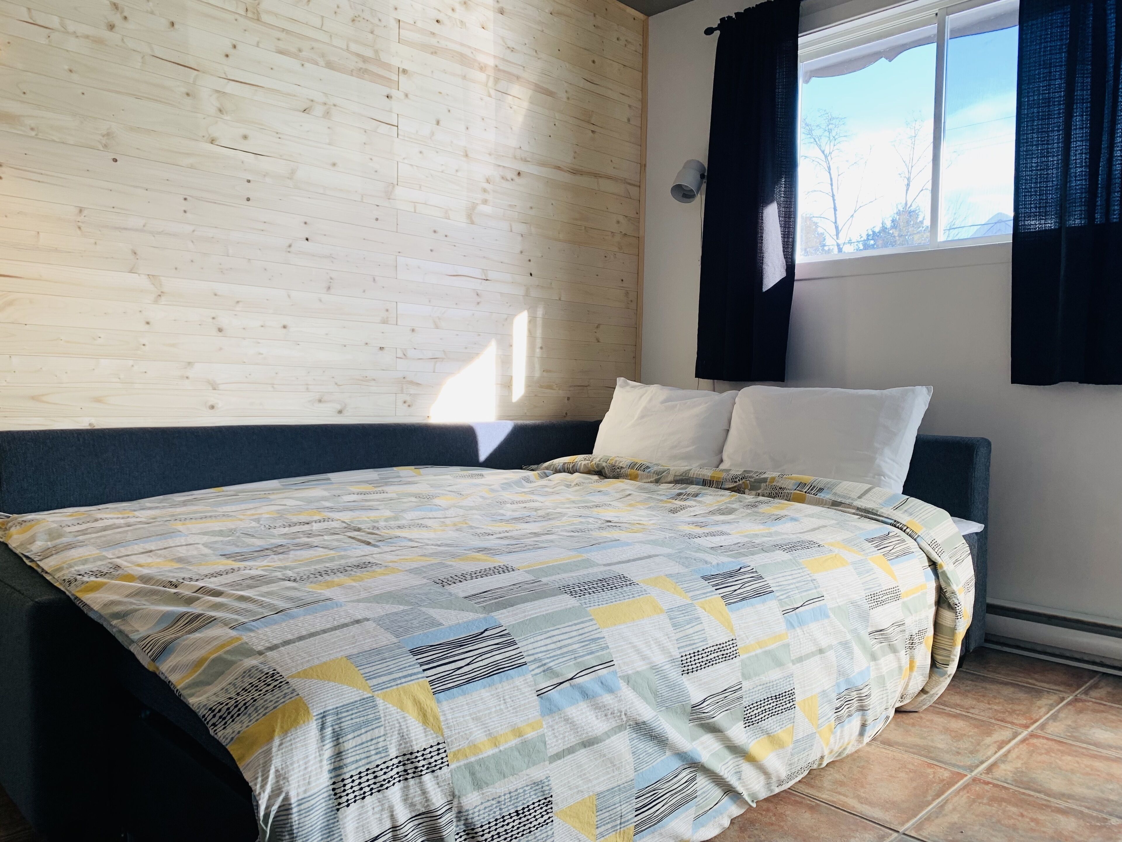 Un dormitorio con pared de madera, una cama con colcha estampada, dos almohadas, una ventana con cortinas negras y una lámpara fijada a la pared.