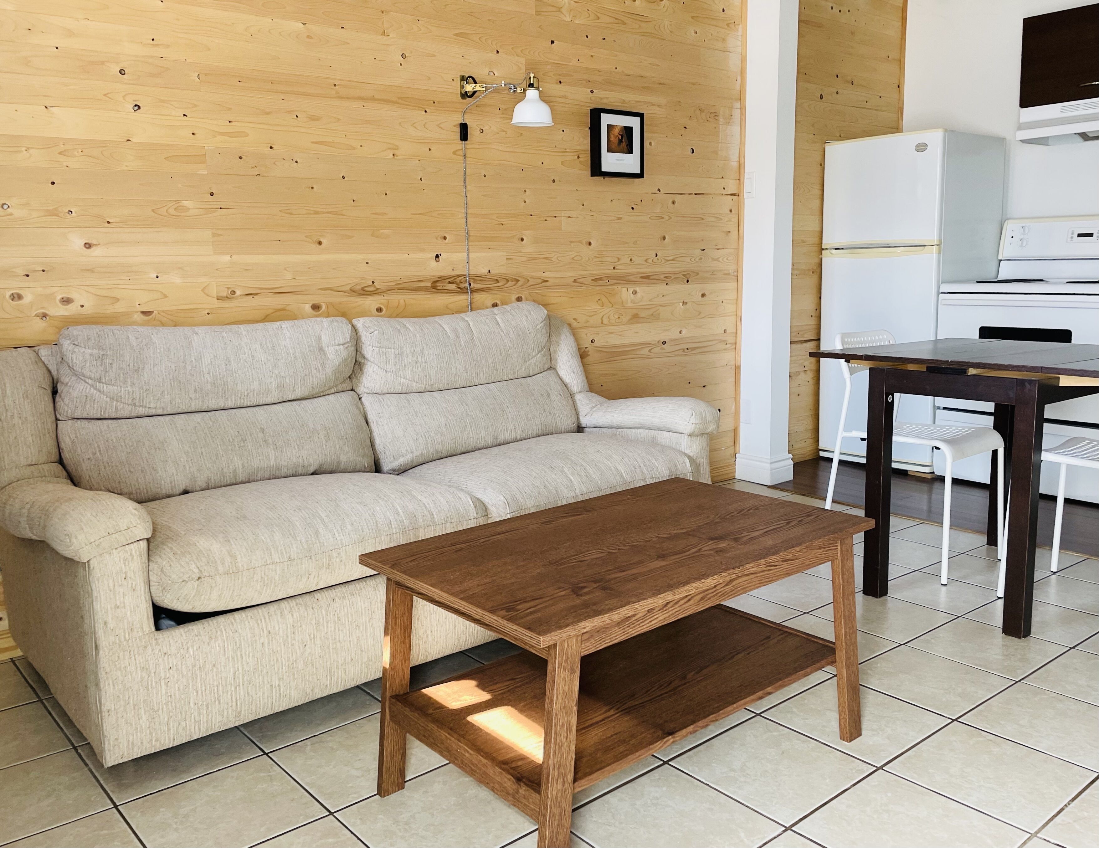 Una pared de madera, un sofá beige, una mesa de centro de madera, un refrigerador blanco y una lavadora blanca.